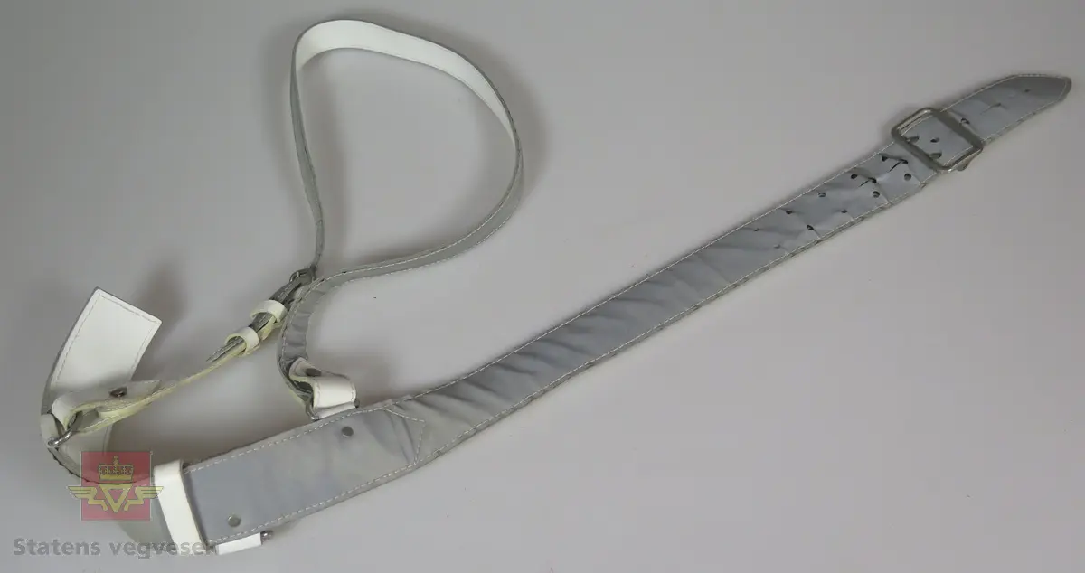 Sølvgrått og hvitt bandolær, med påsydd refleksbånd. Bandolæret reguleres med metallspenne og har regulerbar skulderreim. To festeanordninger i metall.