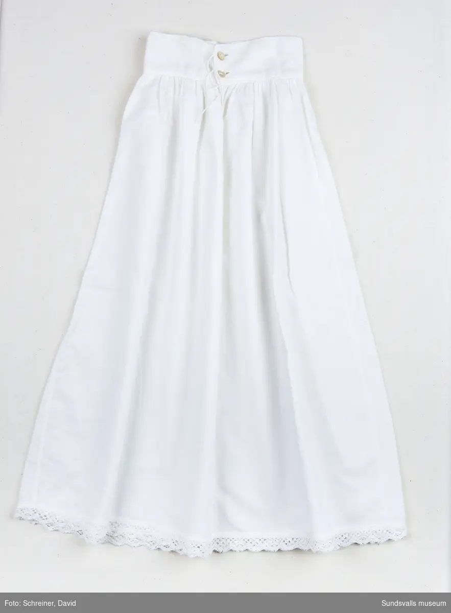 En vit bomullskjol med bred linning som knäpps baktill med två vita knappar. Kjolens nederkant har dekorerats med en brodyrspets.