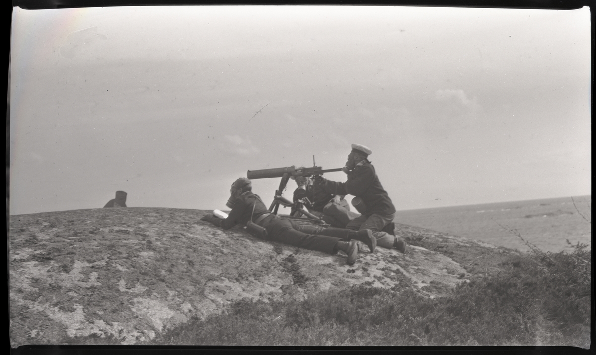 Tre militärer i gasmask bemannar kulspruta på klippa vid havet. Andra bilder i Bergers samling tyder på att detta fotografi tagits vid stridsövningen på Stabbo i Stockholms södra skärgård 1929, detta har dock inte kunnat bekräftas.
