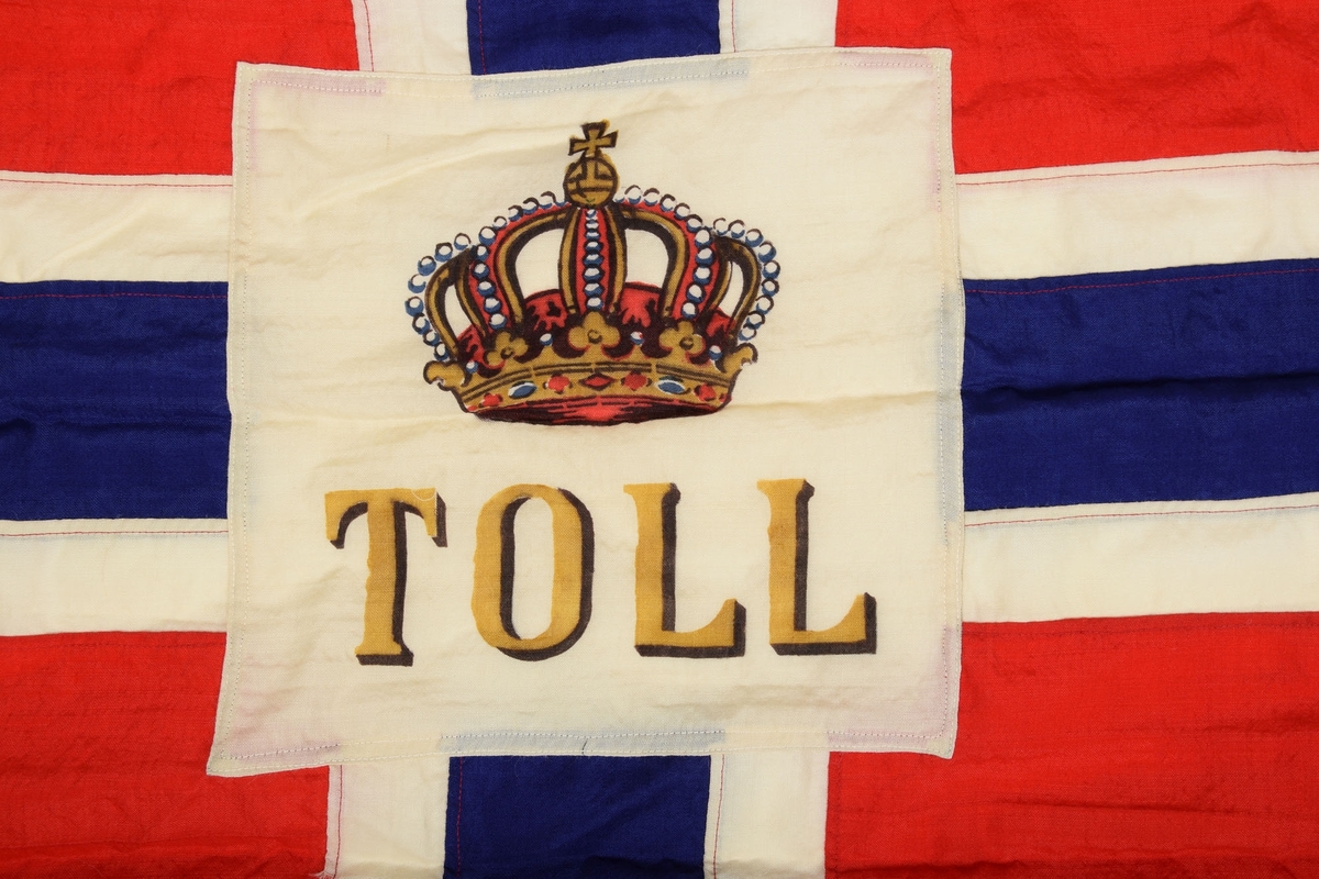 Norgesflagg med "TOLD" skrevet på midten. Over teksten er det avbildet en krone. Bånd i venstre kortside, for oppheng. 3 spisser i den høyre siden.