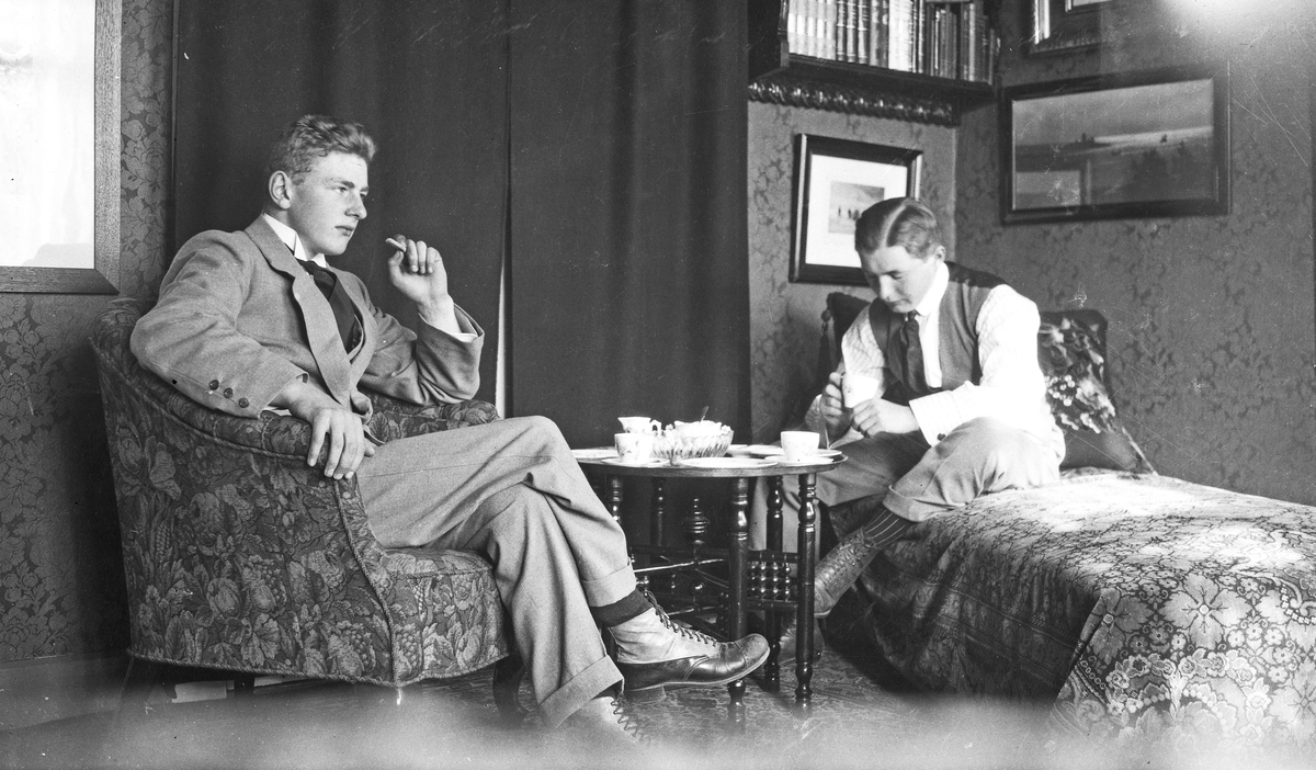 Interiør. To menn sitter og slapper av i stuen. Den ene sitter i en stol og røyker. Den andre sitter på en divan og drikker kaffe. Hverdagsfoto.