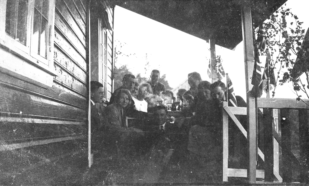 17. mai feiring. Gruppebilde. Tretten personer, seks kvinner og syv menn sitter og røyker på en overbyggd flaggpyntet veranda. Hverdagsbilde.