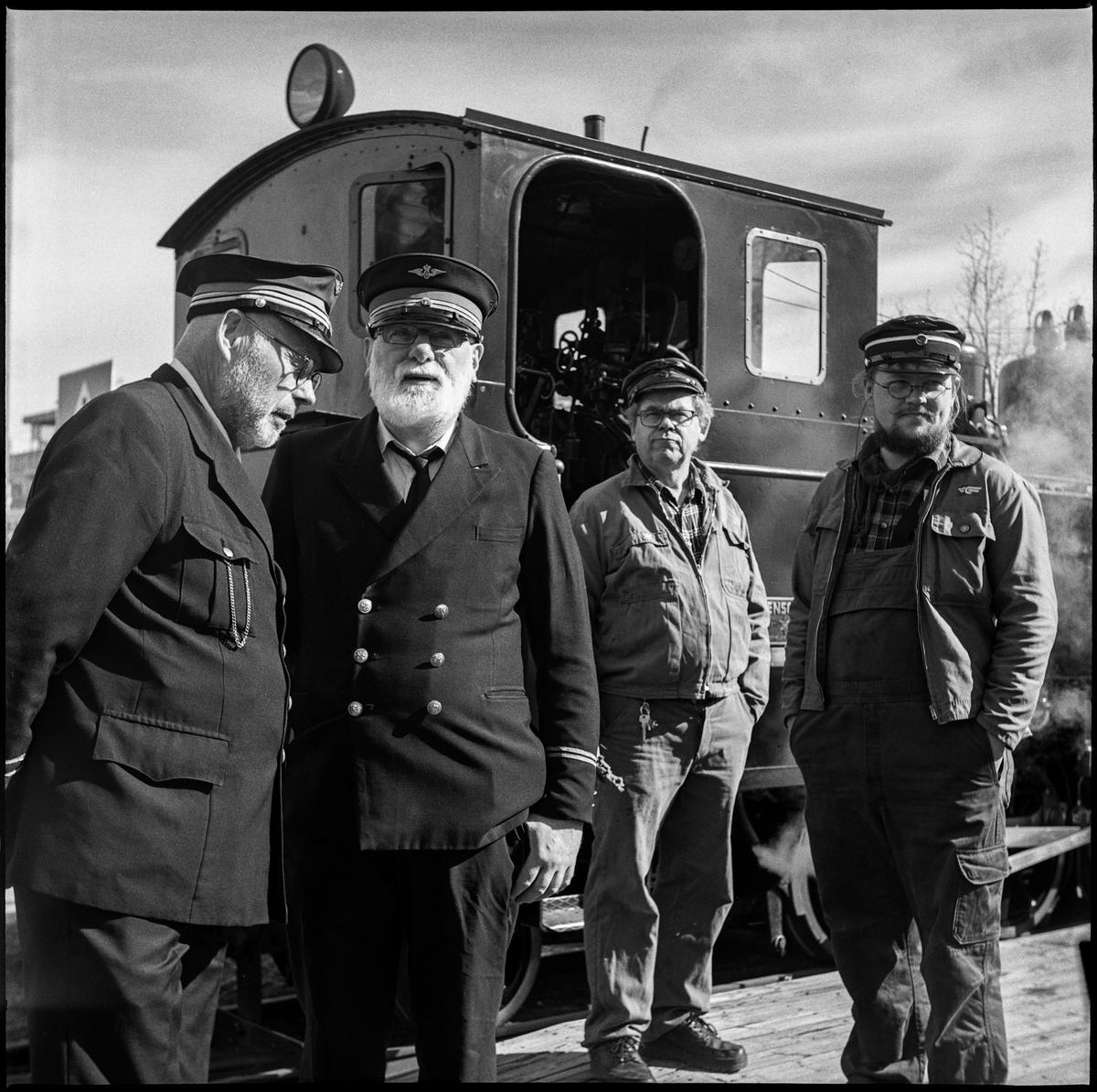 Fire mannlige frivillige i tjeneste.  To av de frivillige er i uniform. Mennene står foran et damplokomotiv.