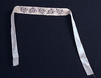 Brudstrumpeband av ljust rött siden med broderier av pärlor i svart och vitt samt silke i svart bestående av fem buketter.
Kantat med vit snodd av sammet, baksidan av vitt siden.