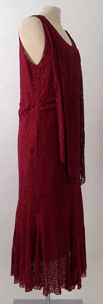 Lång klänning av röd silkesspets med underklänning av rött siden. Ett långt band i röd silkesspets är fastsytt vid vardera axel framtill på klänningen.