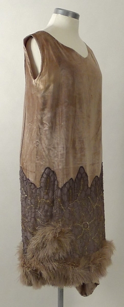 Klänning av ljusgrå silkesammet. På kjolen silverspets och längst ner grå plymer. I midjan en bukett tygblommor i rosa och blått. 1920-talet