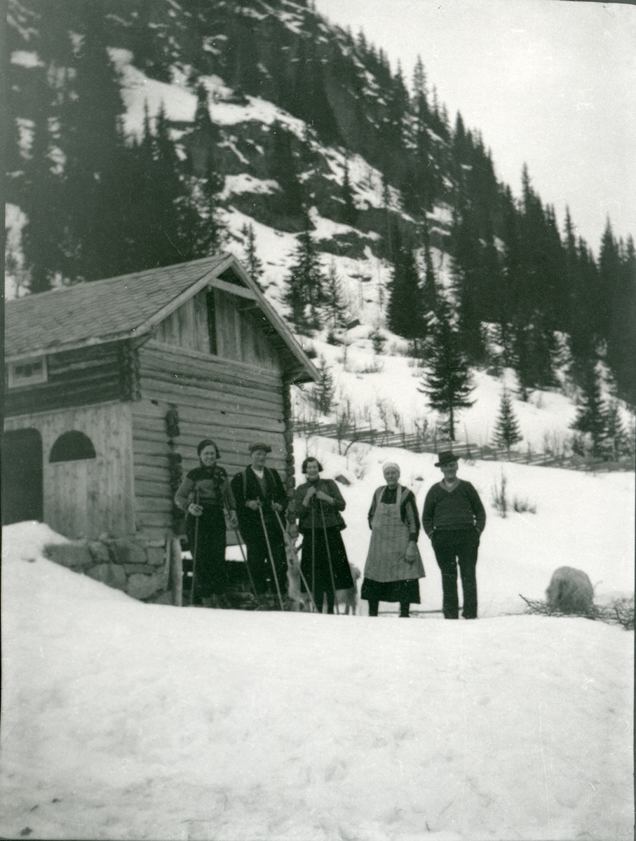 5 personer står foran en tømmerlåve, og 3 av dem har skiutstyr