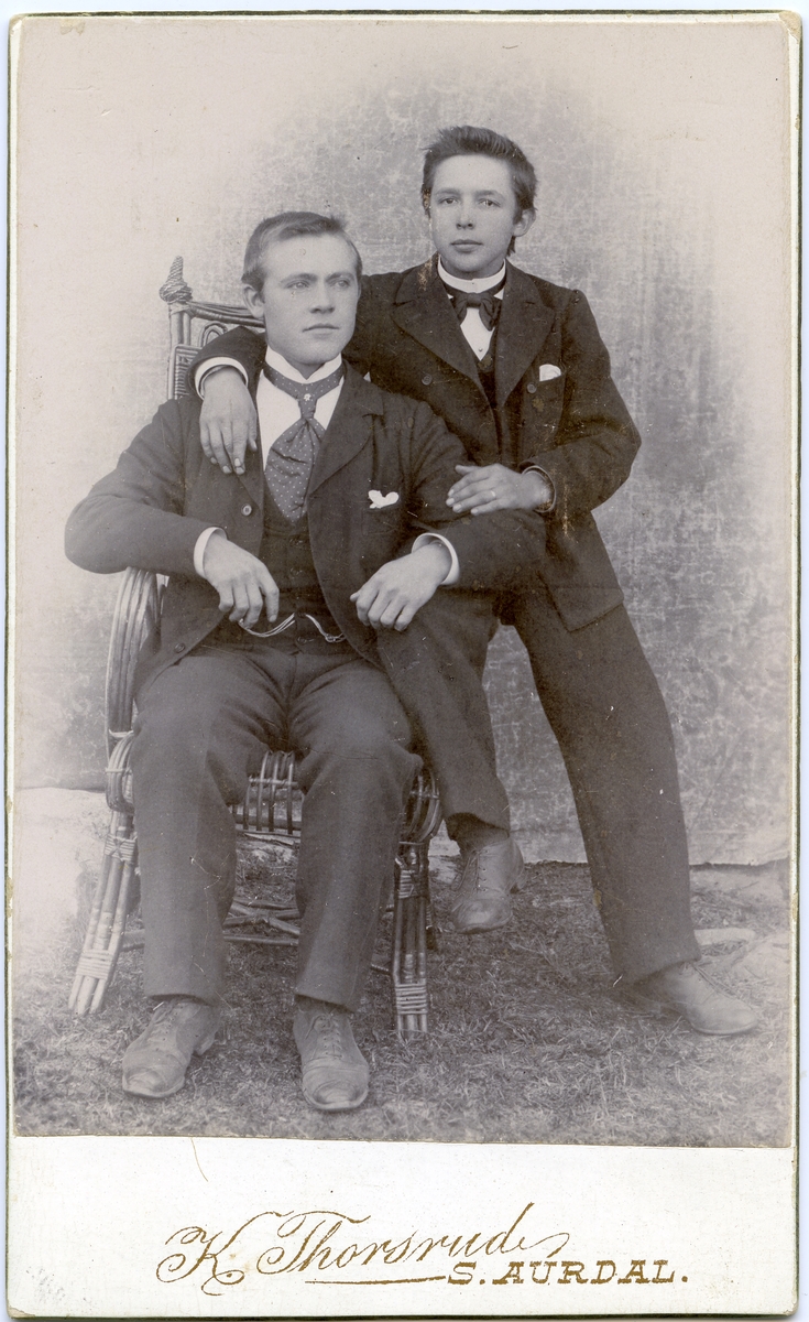 Portrett i helfigur av to unge menn. Den ene sitter i en kurvstol og den andre sitter på armlenet.