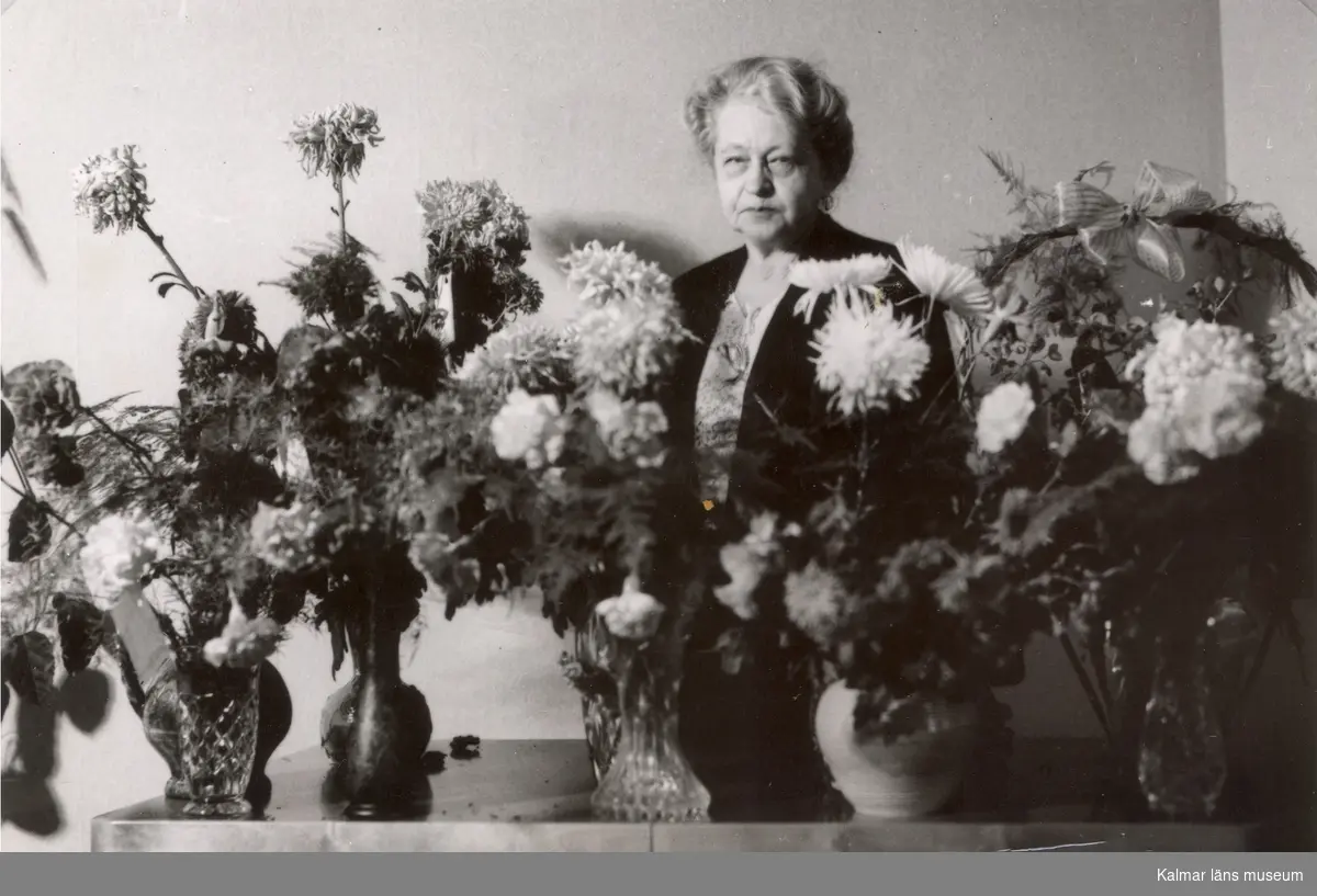 Ellen Warholm stående bakom ett bord fullt med blomsterkvastar, troligen vid någon högtidsdag.
