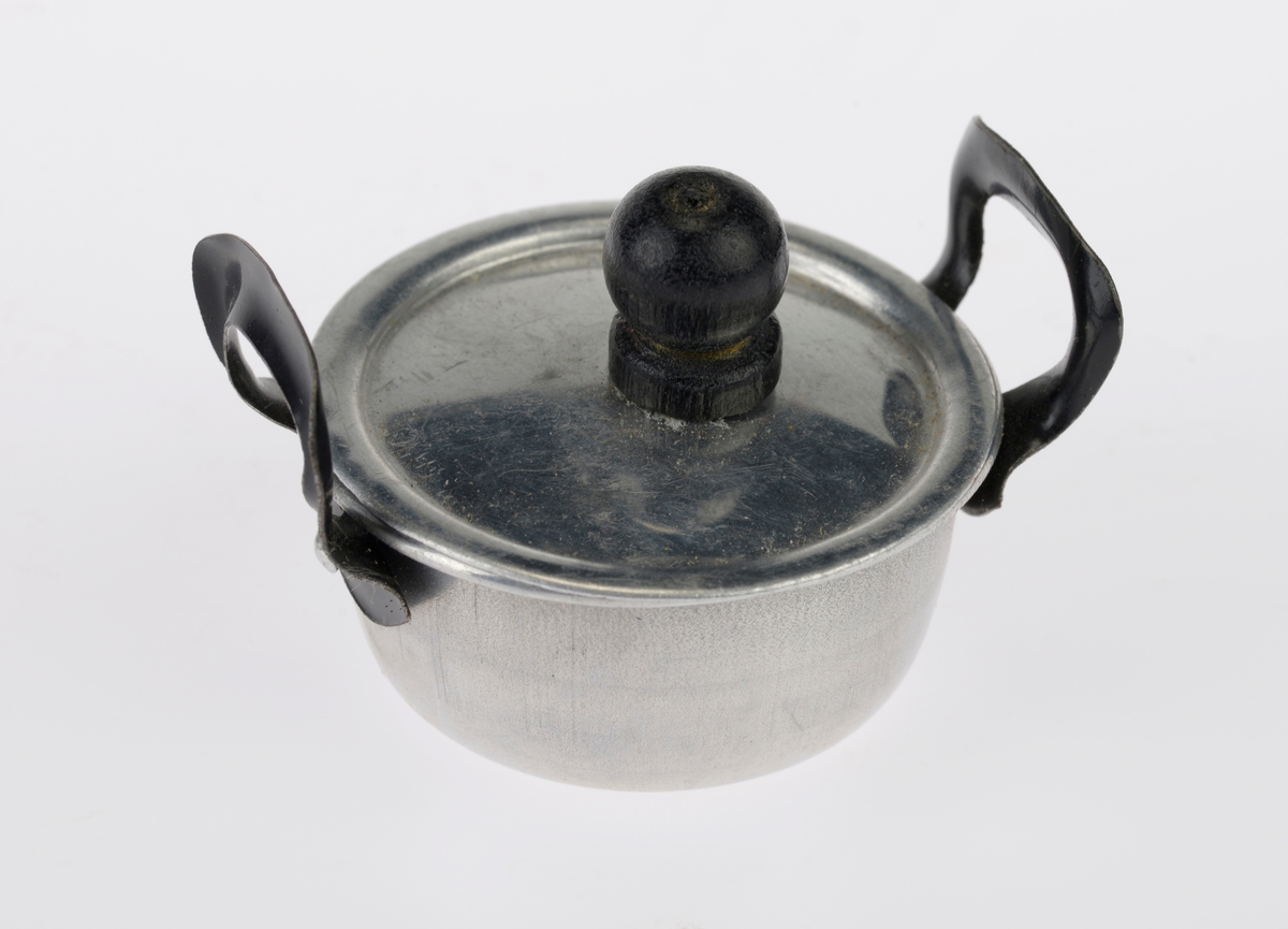 Et kjøkkensett som er montert i en liten pappeske med lokk. Settet er trolig fabrikkprodusert. Gjenstandene er undernummerert 01-10.
FTT.54457.01: En kasserolle med lokk laget av blikk. I hver side av kasserollen er det relativt store svartmalte håndtak av metall, og på lokket er det en treknott som også er svartmalt. Håndtakene og knotten er loddet på. Kasserollen er trolig valset.
FTT.54457.02-03: To identiske tallerkener laget av blikk. De er runde med en bred rand.
FTT.54457.04-05: To identiske kniver laget av metall, og de er antageligvis støpt. Håndtaket har prikker langs kanten som står opp fra metallet, og i midten er det en opphøyet stripe av metall. Knivbladet er relativt bredt og avrundet i tuppen. 
FTT.54457.06-07: To identiske skjeer laget av metall, og de er antageligvis støpt. Håndtaket har prikker langs kanten som står opp fra metallet, og i midten er det en opphøyet stripe av metall. 
FTT.54457.08-09: To identiske gafler laget av metall, og de er antageligvis støpt. Håndtaket har prikker langs kanten som står opp fra metallet, og i midten er det en opphøyet stripe av metall. Gaflene fire tenner.
FTT.54457.10: Esken der gjenstandene 01-09 er montert i. Esken og lokket er laget av papp og er stiftet i hjørnene. Esken er grå og blå på farge. Langs kanten inni esken er det klippet ute en tungekant. I bunnen er det en rød papplate der det er klippet ut hull for montering av de ulike gjenstandene.