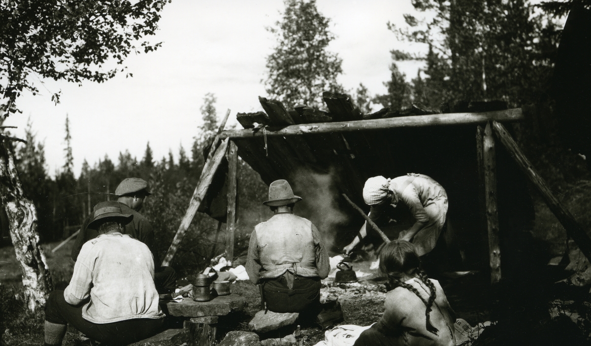 Kafferast ved "gapahuk" i Galåsen (?). Tre menn og ei jente sitter med ryggen til. Kvinne koker kaffe.