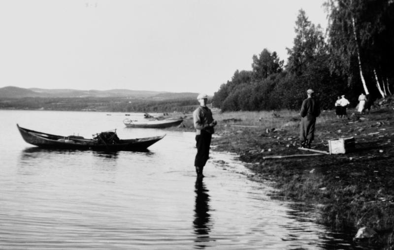 Svart-hvitt foto av en tom åfløy (robåt) som ligger påe t stille vann, en mann står uti vannet og snakker med en mann som sitter på land.