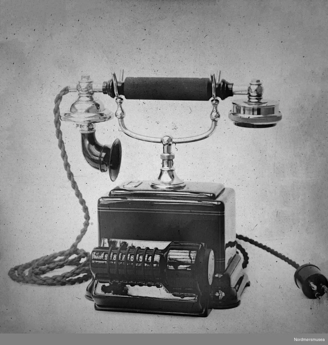 Foto av en eldre telefon fra Kristiansund. sjelden type nummerskive. Telefonen kan dateres til etter 1923. 
Telefonen i Kristiansund ble automatisert i 1923, tidligere måtte en nytte sveiv for å kalle opp sentralen, opplyser Kjell R. Johansen 2018.
Fra Nordmøre museums fotosamlinger.