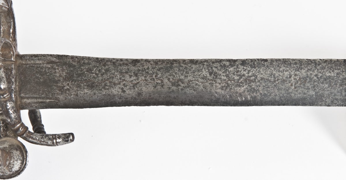 Soppformet knapp med nitteknapp Oversiden av knappen har skåret og meislet dekor. Grep mangler .Asymmetrisk  feste av runde bøyler Sidering på utsiden som ender i en knapp. Sideringen på innsiden er borte. Klingebrekker på innsiden. Håndbøylen er brukket rett under knappen Sidebøyle og tersbøyle. Bøylene har smidd og meislet dekor. Flere bøyler på innsiden er brukket.
Rett tveegget klinge med ovalt tverrsnitt  Klingen merket mes en snal bølgelinje innlagt i kobber.