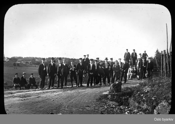 En gruppe menn og ungdommer poserer for fotografen i et veikryss, 5. juli 1903. Haslum kirke bak til venstre. Haslum gård.