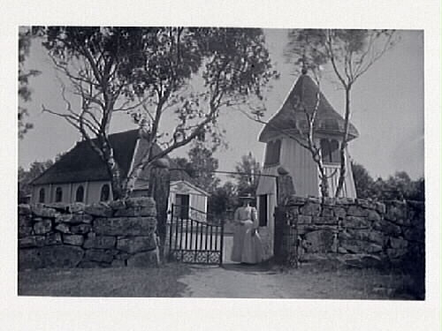 Nösslinge kyrka och klockstapel. En kvinna står vid ingången till kyrkogården.