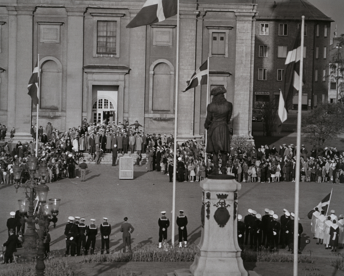 Militärer och civila har samlats på Stortorget. En grupp civilklädda män med vita mössor står på Fredrikskyrkans trappa. I bakgrunden bakom kyrkan skymtar militärhemmet.