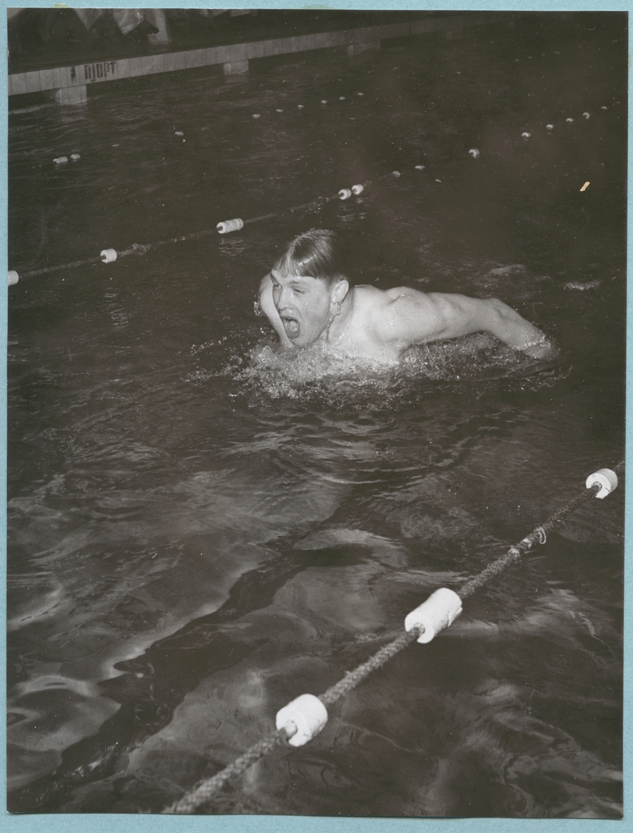 En manlig simmare syns ovanför ytan mitt i ett simtag. Han befinner sig mitt i en bana i en simbassäng. Fototo är daterat till 15-3-52.