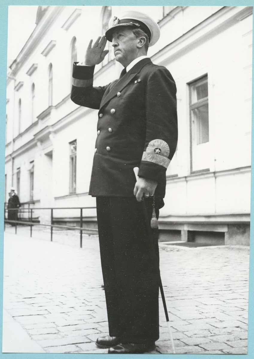 Konteramiral Samuelson, klädd i uniform, står utanför bataljon Sparres kanslibyggnad och gör honör.