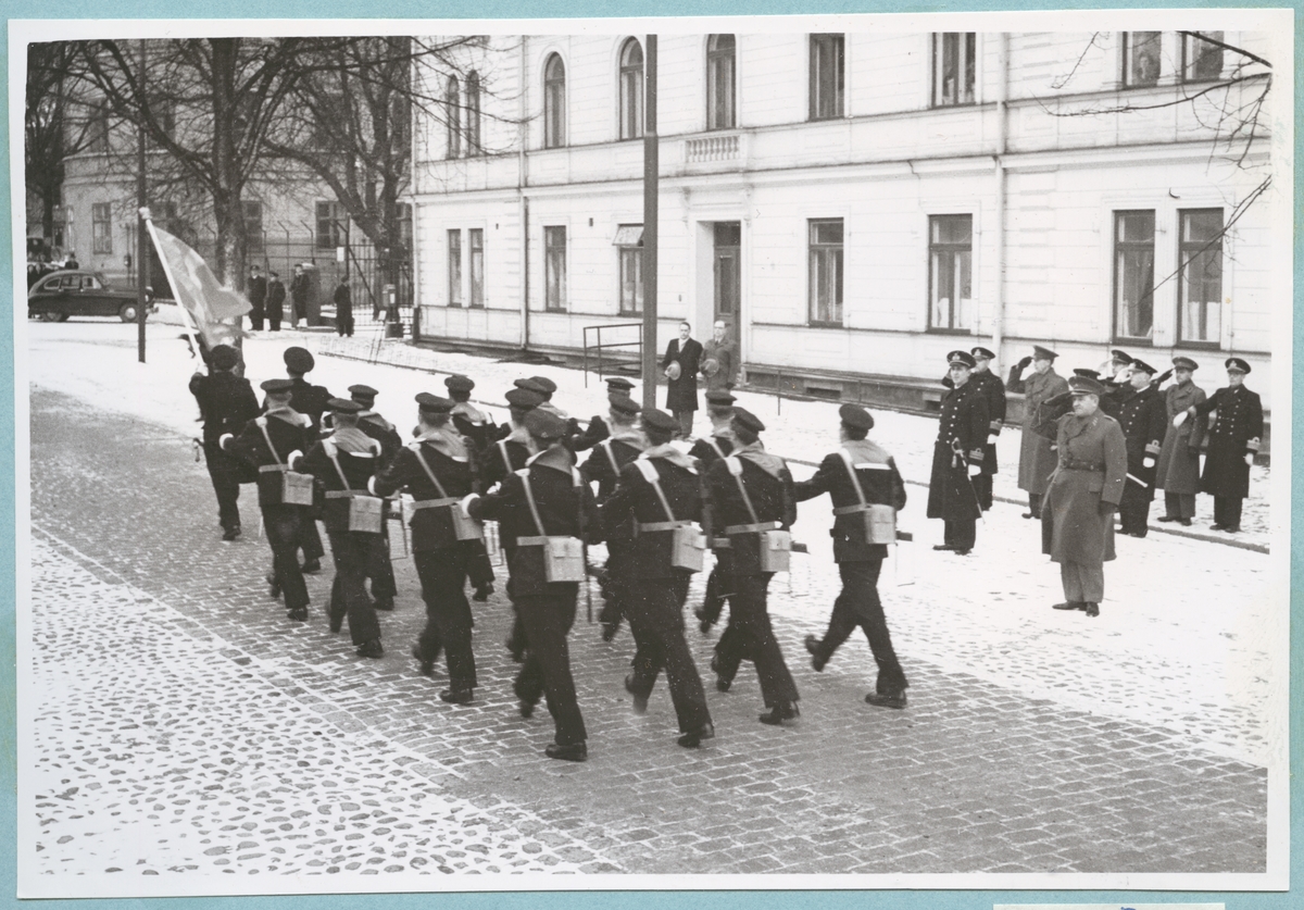 En grupp uniformerade flottister marscherar i samlad trupp längs med Drottninggatan. Främst går en militär som håller upp en svensk flagga. På den gat- och kullerstensbelagda gatan ligger ett tunt snötäcke. Vid sidan om de marscherande flottisterna står officerare och gör honnör framför Bataljon Sparre. I bakgrunden syns även några civila åskådare.