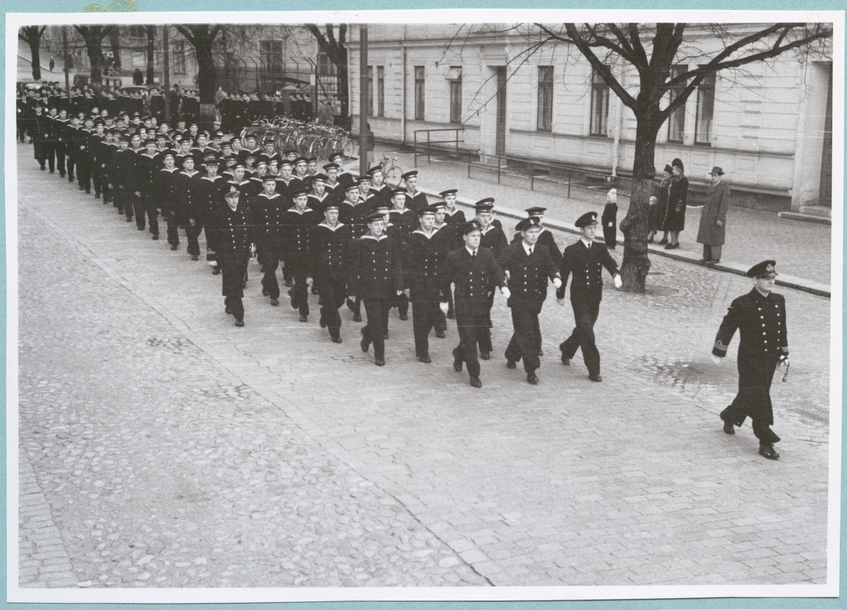 Uniformsklädda flottister marscherar längs med Drottninggatan under ledning av en kapten. Den stenlagda gatan kantas av bataljon Sparres kanslibyggnad. På gatan står även ett par civila åskådare. 
Kyrkparad för nyinryckta värnpliktiga.