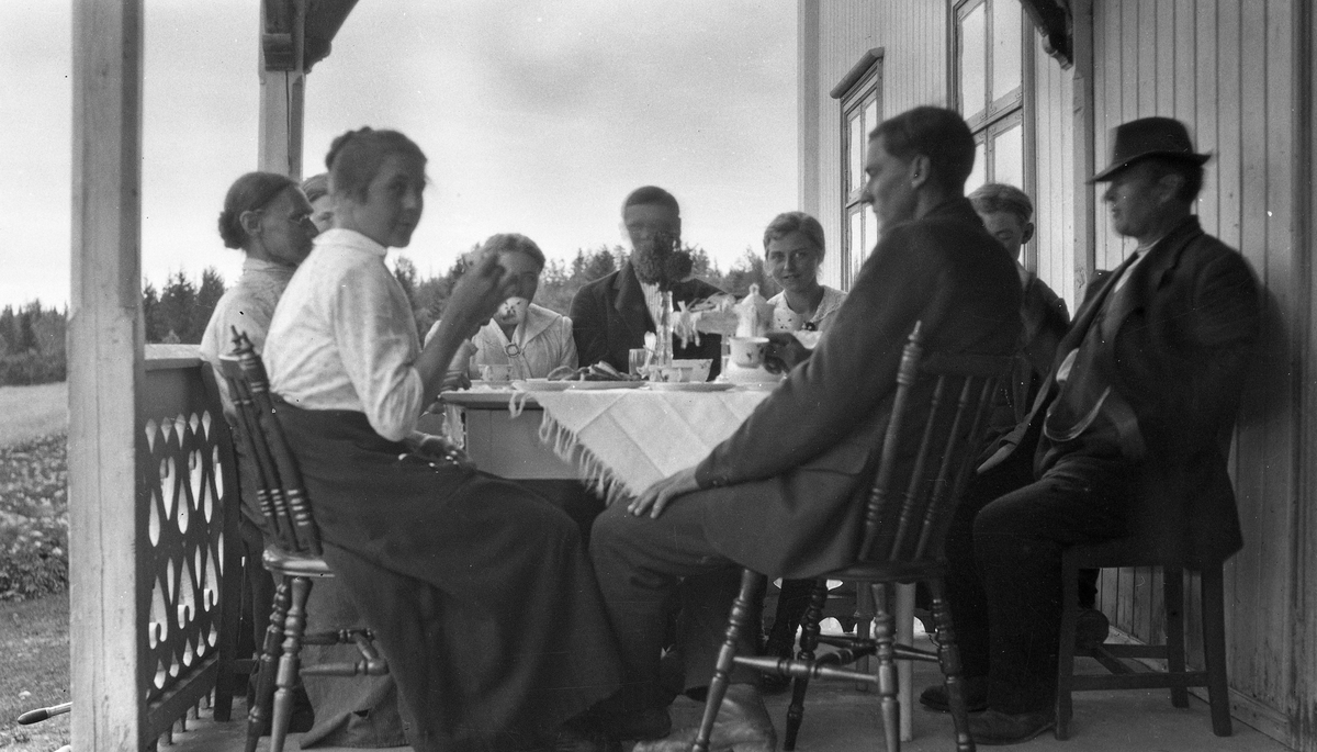 Personer samlet rundt et kaffebord på en veranda. Bak bordeet med front mot fotografen sitter til venstre Anna Tømta (f1904), til høyre hennes søster Sigrid Tømta (f.1900). Nr. to fra høyre, som man bare ser litt av hodet på, er muligens deres bror Paulus Tømta (f.1899). Bildet som, er fra ca. 1920, er sannsynligvis fra Skullerud i Rossgutua på Skreia, som var Tømta-familiens hjemsted.
