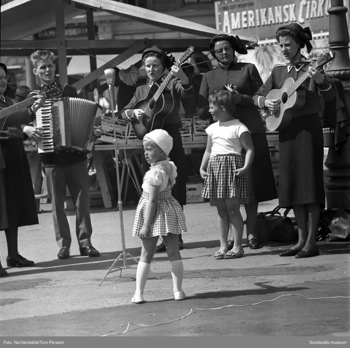 Frälsningsarmén spelar och sjunger på torget i samband med Barnens Dag. Två små flickor (systrar?) ser ut att vilja delta i uppträdandet.
