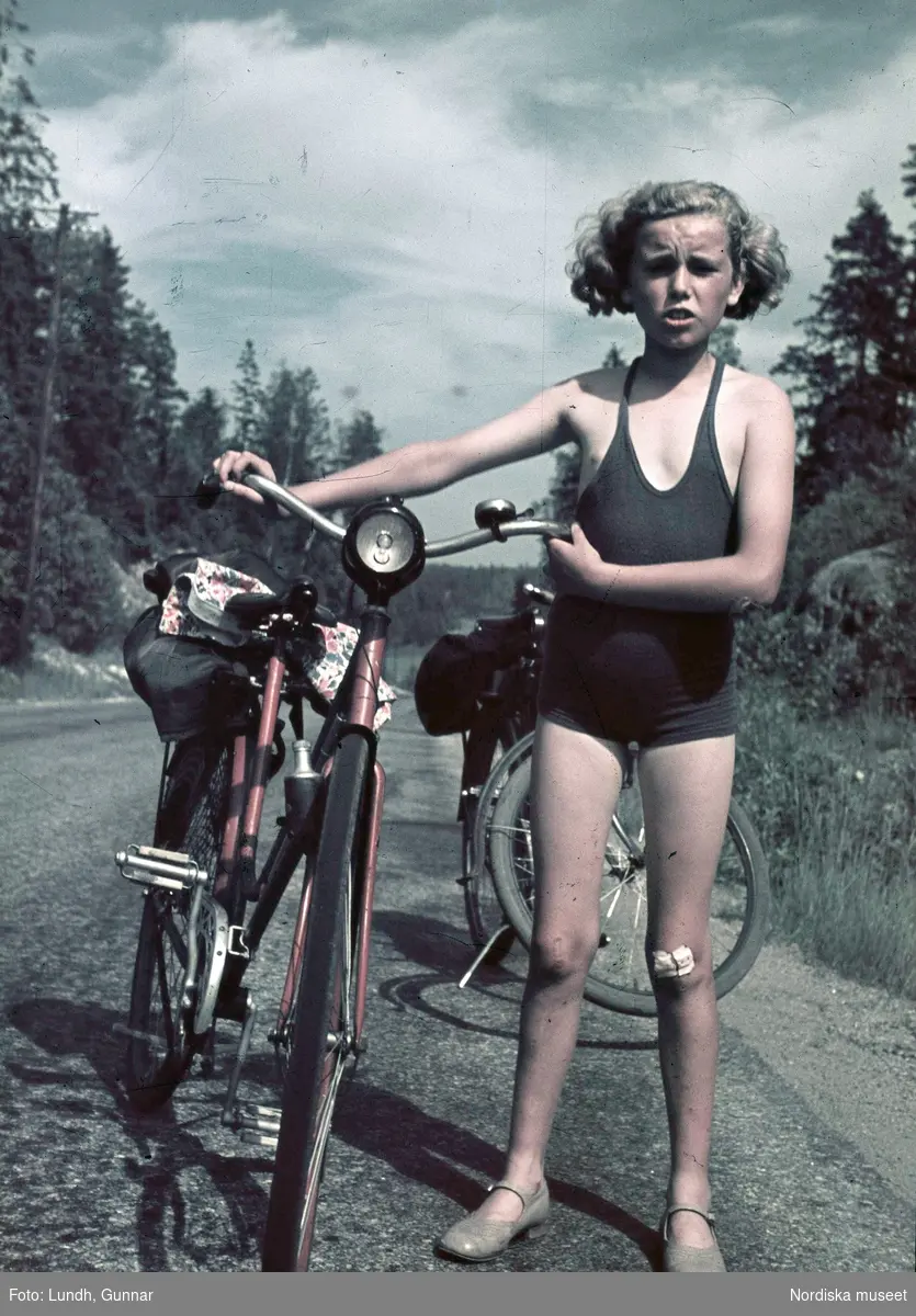 Flicka, fotografens dotter Jytte, i blå baddräkt med cykel. På vänster knä ett plåster. Troligen fotograf Gunnar Lundhs dotter Jytte.