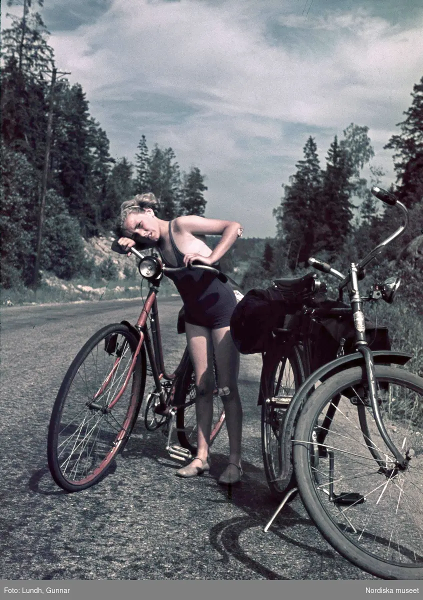 Flicka i blå baddräkt vid två cyklar. På vänster knä och armbåge har hon plåster. Troligen fotograf Gunnar Lundhs dotter Jytte.