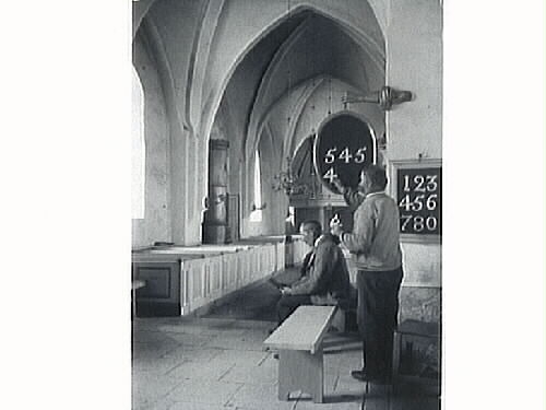 Två män i en kyrka. Den ene är troligen kyrkvärd och sätter upp psalmnumren, den andre sitter bredvid på en bänk och läser. I bakgrunden syns en stor kamin. Inga uppgifter om kyrka eller personer.