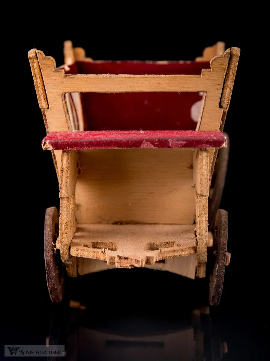 En liten leke- hestevogn i kryssfiner. To store og to små hjul, kledd med rød filt og vokspapir er brukt som vinduer.