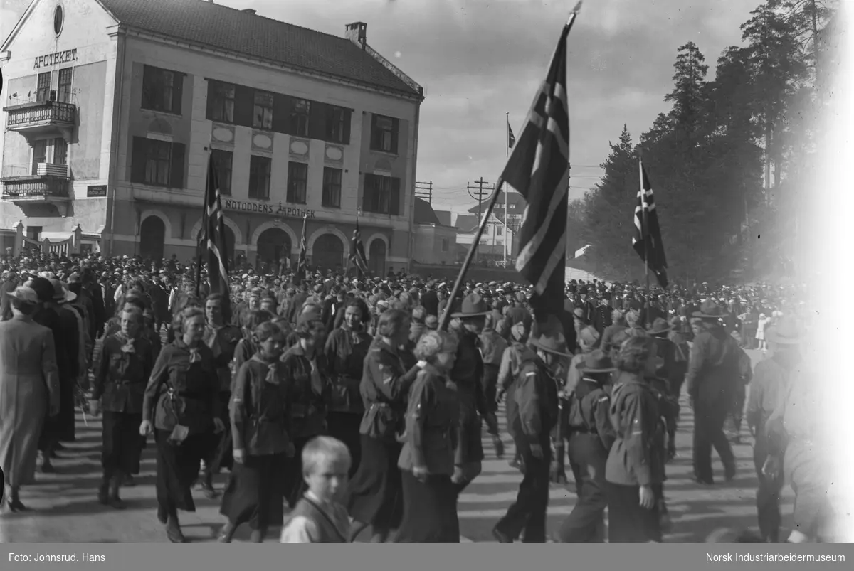 17. mai 1935. Speidere marsjerer med store flagg på torget. Folkemengde lags Notodden apotek.
