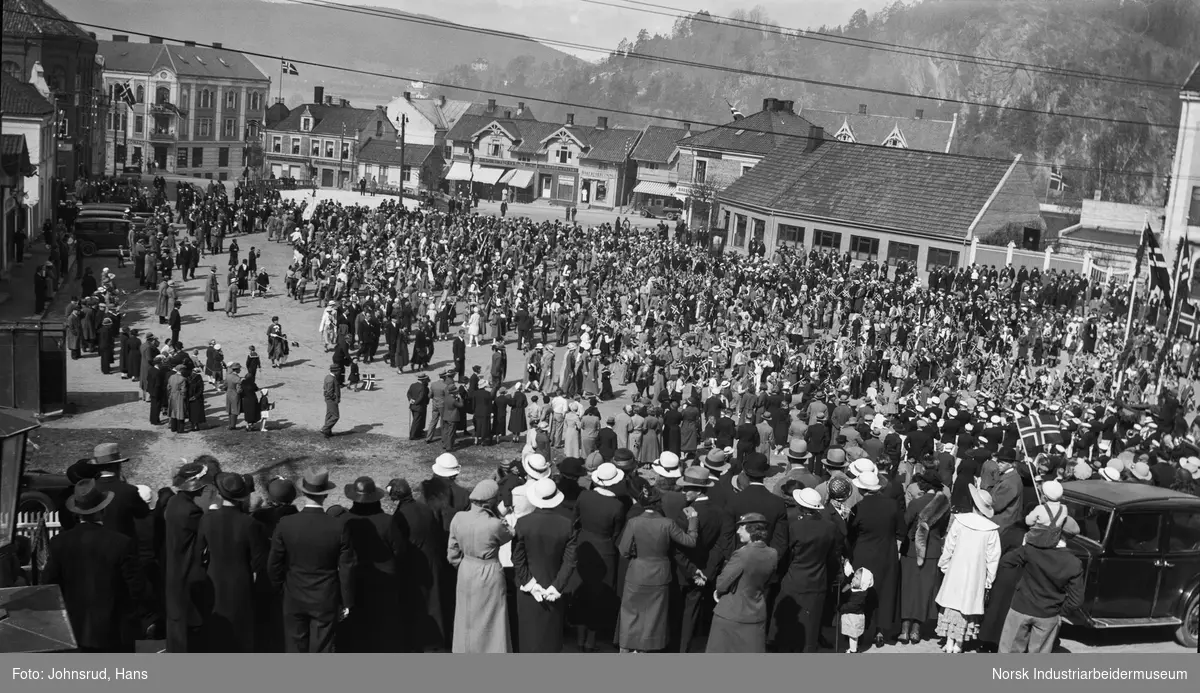 17. mai 1933. Folkemengde med flagg samlet på torget. Notodden apotek og øvrige butikklokaler i bakgrunnen.