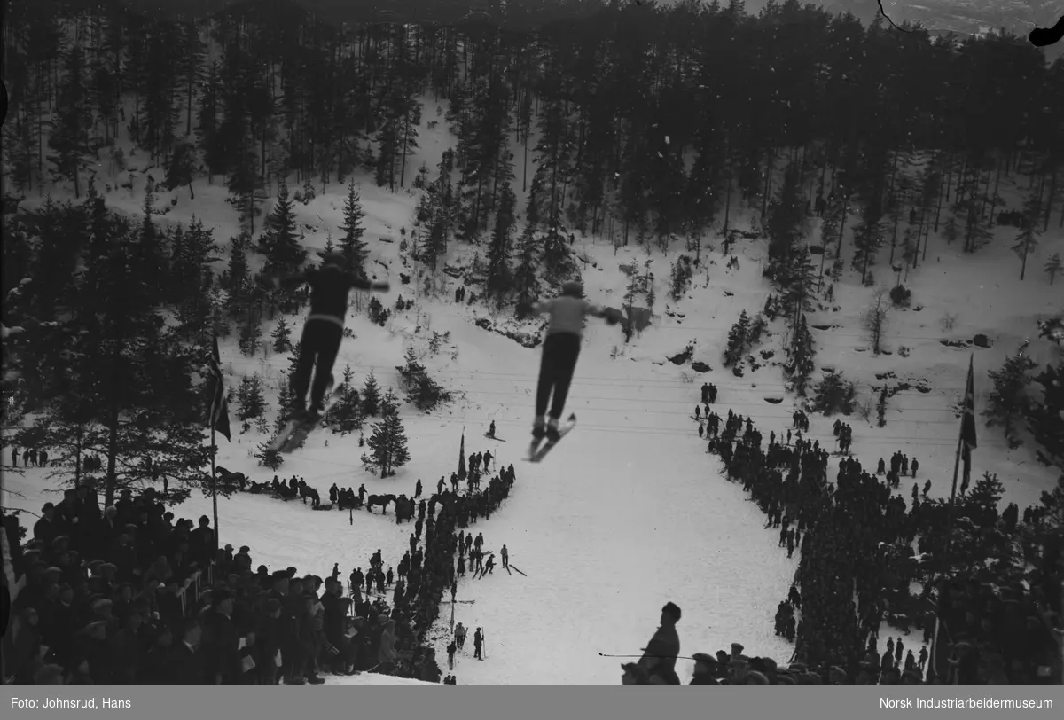 To menn hopper på ski samtidig i Dalsbakken (mellom Eikeskar og Dalsåsen). Publikum står tett sammen nedover hele bakken, noen med norske flagg.