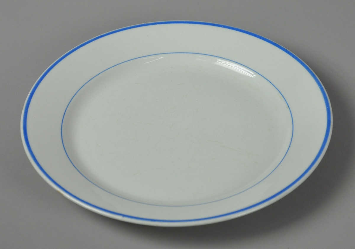Hvit tallerken av glassert keramikk, med to blå striper ved inner- og ytterkant på tallerkenen.
