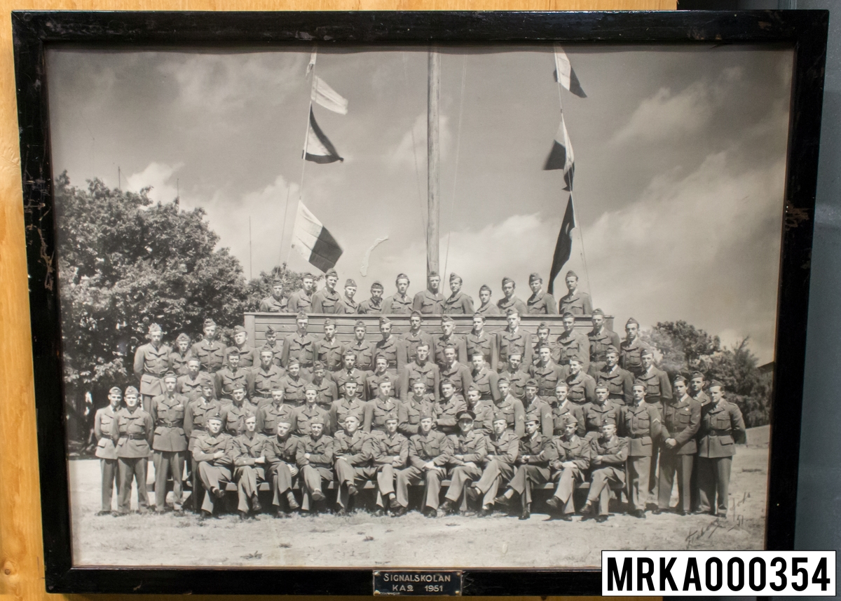 Fotografi taget på befäl och soldater som genomfört grundläggande soldatutbildning på 1:a Batteriet KA 2.
Fotografiet taget utanför gymnastiksalen Gräsvik KA 2.
Flobergs Foto 1951.