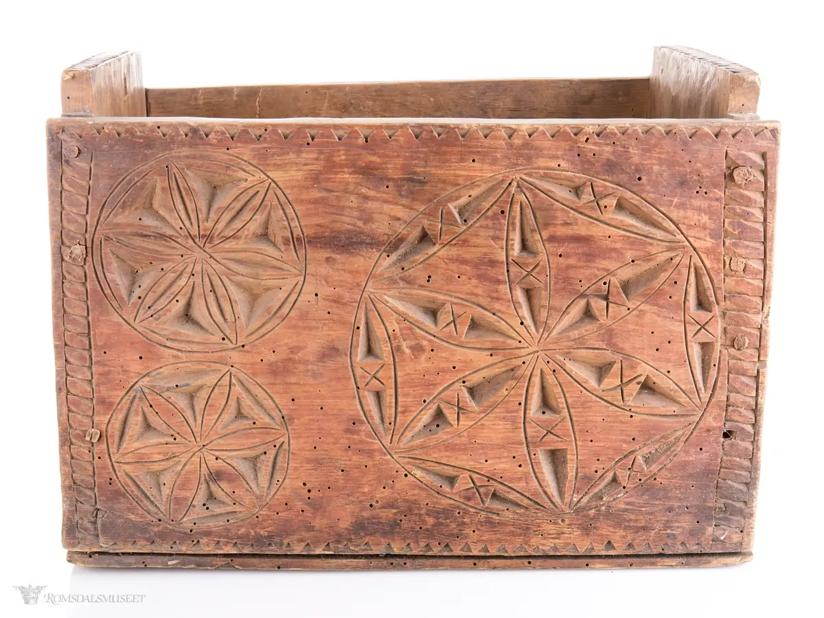 En stor og to små seksbladsroser i karveskurd på langsidene med skraverte dekorfelter og spetter rundt. På kortsidene er det skåret ut en stor seksbladsroser med rike dekorasjonsfelter bestående av spetter, streker diamantformer, sirkler med strek gjennom og solkors/kors. De største rosenen har kryss skåret inn på kronbladene.