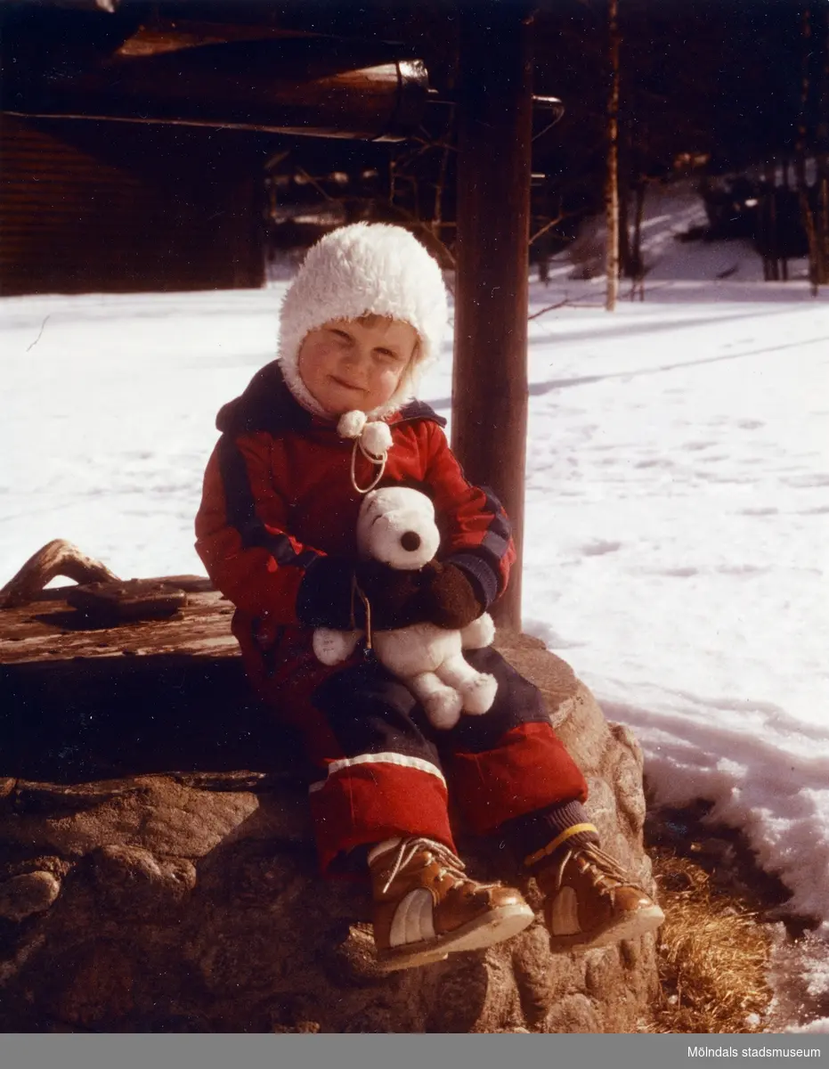 Lilla Karin sitter utomhus på ett brunnslock. I famnen har hon ett gosedjur (Snobben). I landskapet ligger snö och familjen är med bekanta på påskutflykt till Västerdalälven i Dalarna, 4-6 april 1979.