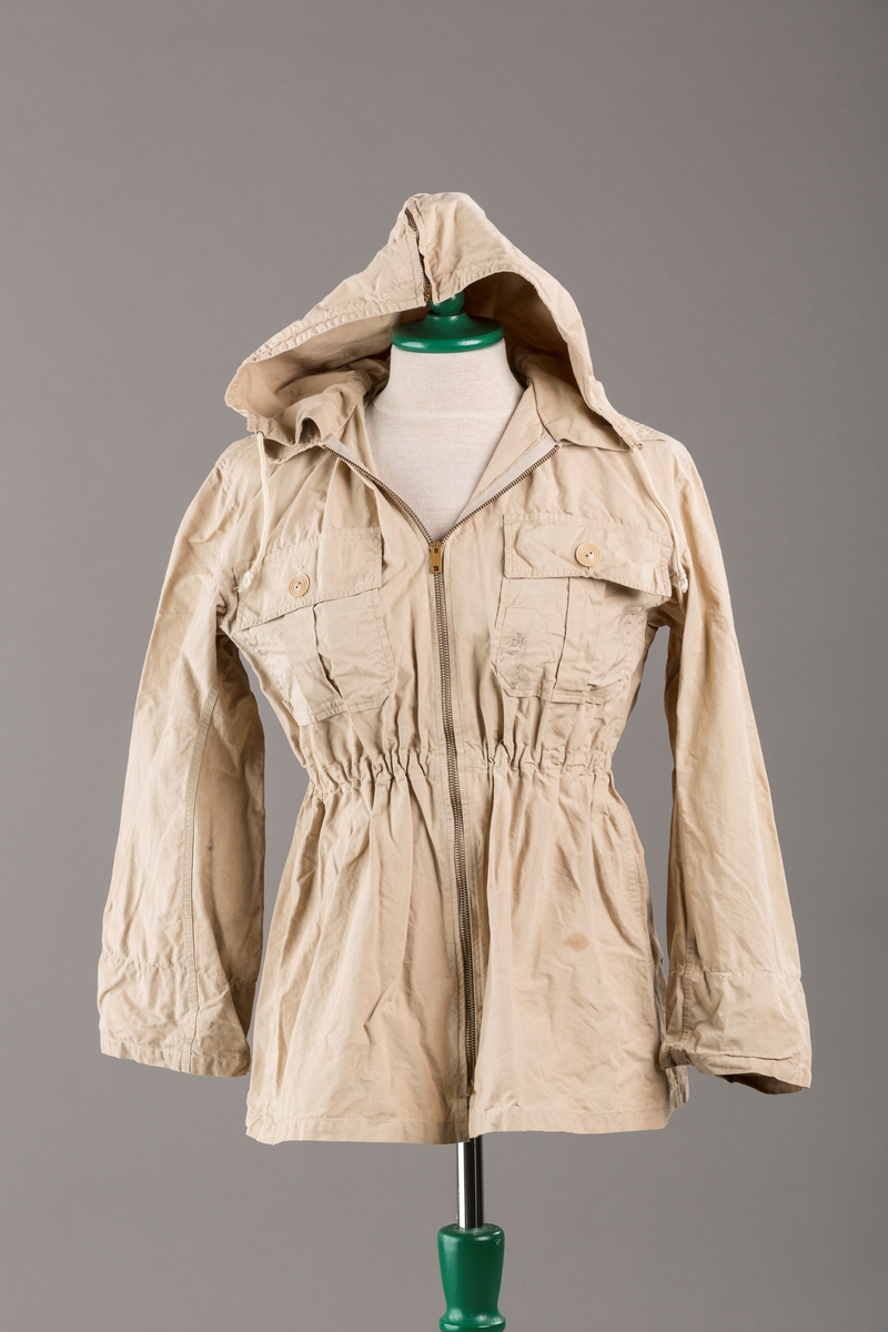 Form: Vanlig anorakkfasong med strikk i livet. 2 brystlommer med knapp.
Hette med glidelås på topp midt, snøring.
Glidelås helt ned foran.
Innermansjett med strikk i ermene.