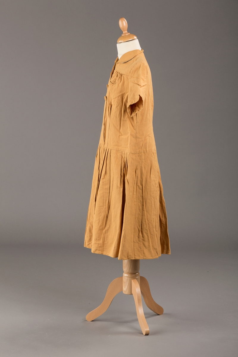 Form: Korte ermer, livet rynket til bærestykke foran, plissert skjørt, tre pynteknapper, en trykknapp, forlomme.