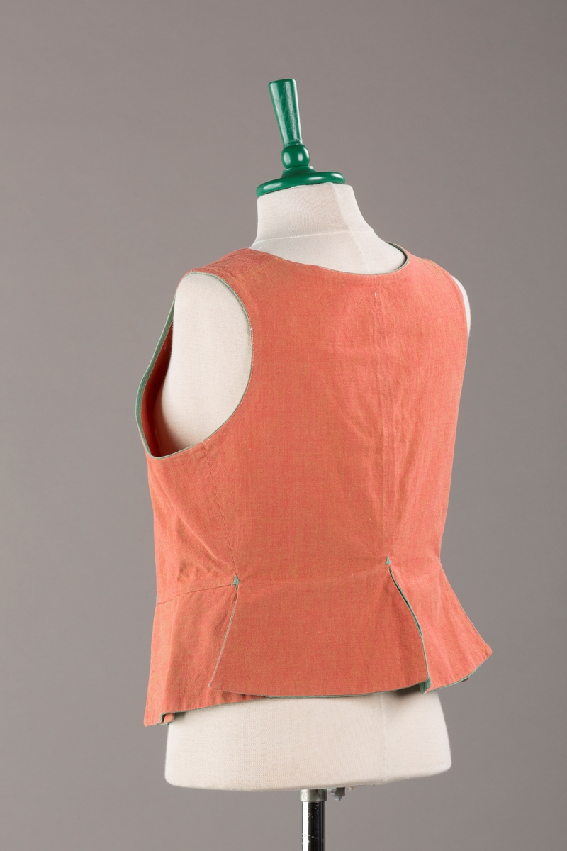 Rustrødt liv med grønn kant og fór, seks skjøter. Håndsydd forsterkning i kontrastfarge øverst i skjøtesplittene. Lukkes med tinnhekte og silkesnor.