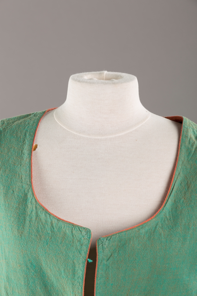 Grønt liv med rustrød kant og fór, seks skjøter. Håndsydd forsterkning i kontrastfarge øverst i skjøtesplittene. Lukkes med tinnhekte.