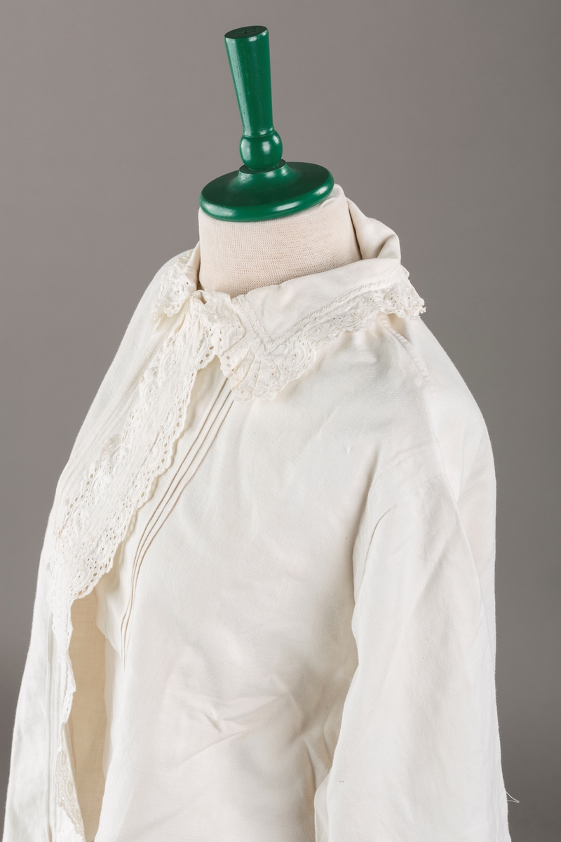 Form: Hoftelang trøye, Lange ermer, krage. Kragen og åpningen foran er kantet med  maskinprodusert blonde. Tre biser på hver side av åpn. foran. Lukkes med to løse knapper.