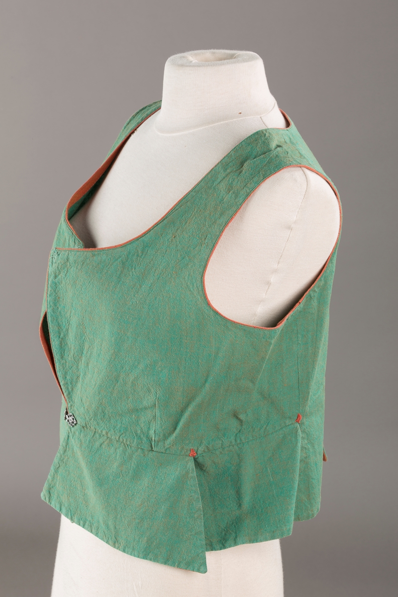 Grønt liv med rustrød kant og fór, seks skjøter. Håndsydd forsterkning i kontrastfarge øverst i skjøtesplittene. Lukkes med tinnhekte.