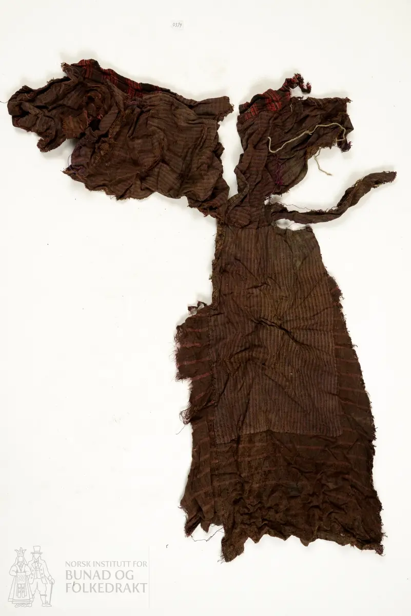 Svært utslitt del av ein stakk med linning, nederkant og ei metallhekte i eine enden av linning. Nedanfor denne delen av linning er det sauma fald som truleg er splitten midt framme på stakken. Av tre ulike verkensstoff, lerretsbinding med bomull i renning, ull i innslag. Stripete stoff i brun og lilla, svært utydeleg lillafargar. Fleire små hol etter mus, har vore nytta som stappefille.