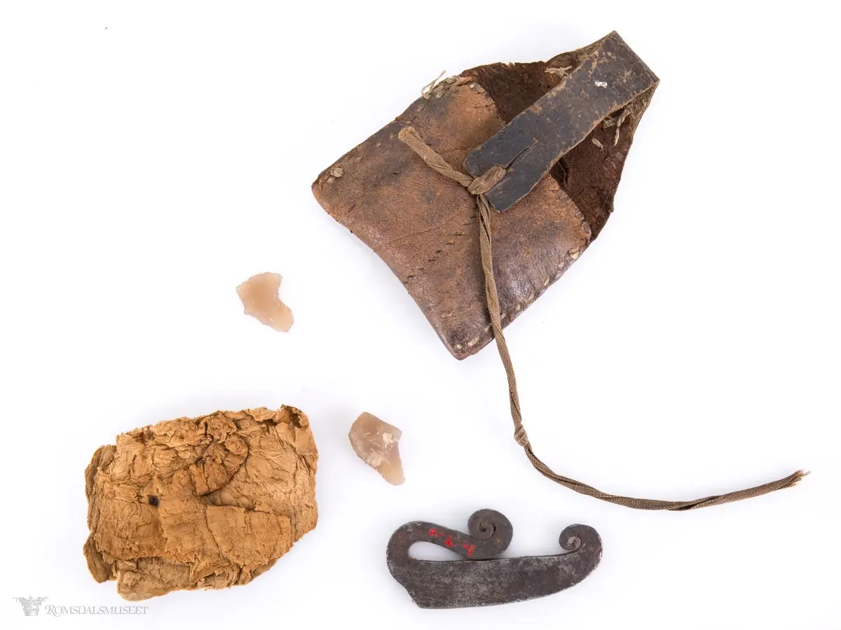 Fyrtøy bestående av en liten lærpose, slagstål, flint og knuskstykker. I posen er det to knuskstykker. Slagstålet har nærmest strykejernsform.