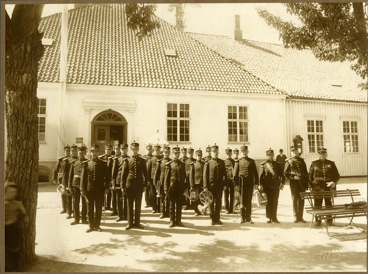 Bildet er av 5.Div Musikkorps i en oppstilling. Bildet tatt etter 1918 (ca 1920) utenfor Kommandantskapets hovedbygning. Munkegata 2, Trondheim.
