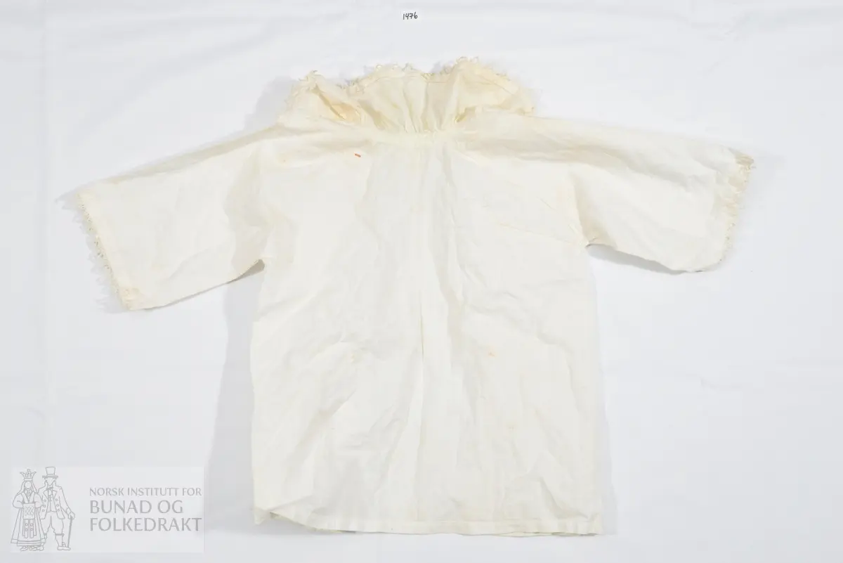 Barneskjorte. Hvitt bomullslerret. Krage med nuperellekant og hullfald er rynket til bolen i små rynker. Løpegang med knytebånd i halsen. Åpen hele veien ned midt foran. Lukkes med knyting i halsen. Ermene er rette og vide, er ikke isydd, men henger fast i ryggen og et bærestykke foran. Denne delen er skåret i ett og sydd til et stykke foran, hvor sømmen er en fortsettelse av sømmen under ermet. Ermene er kantet med nupereller. Kun maskinsøm.