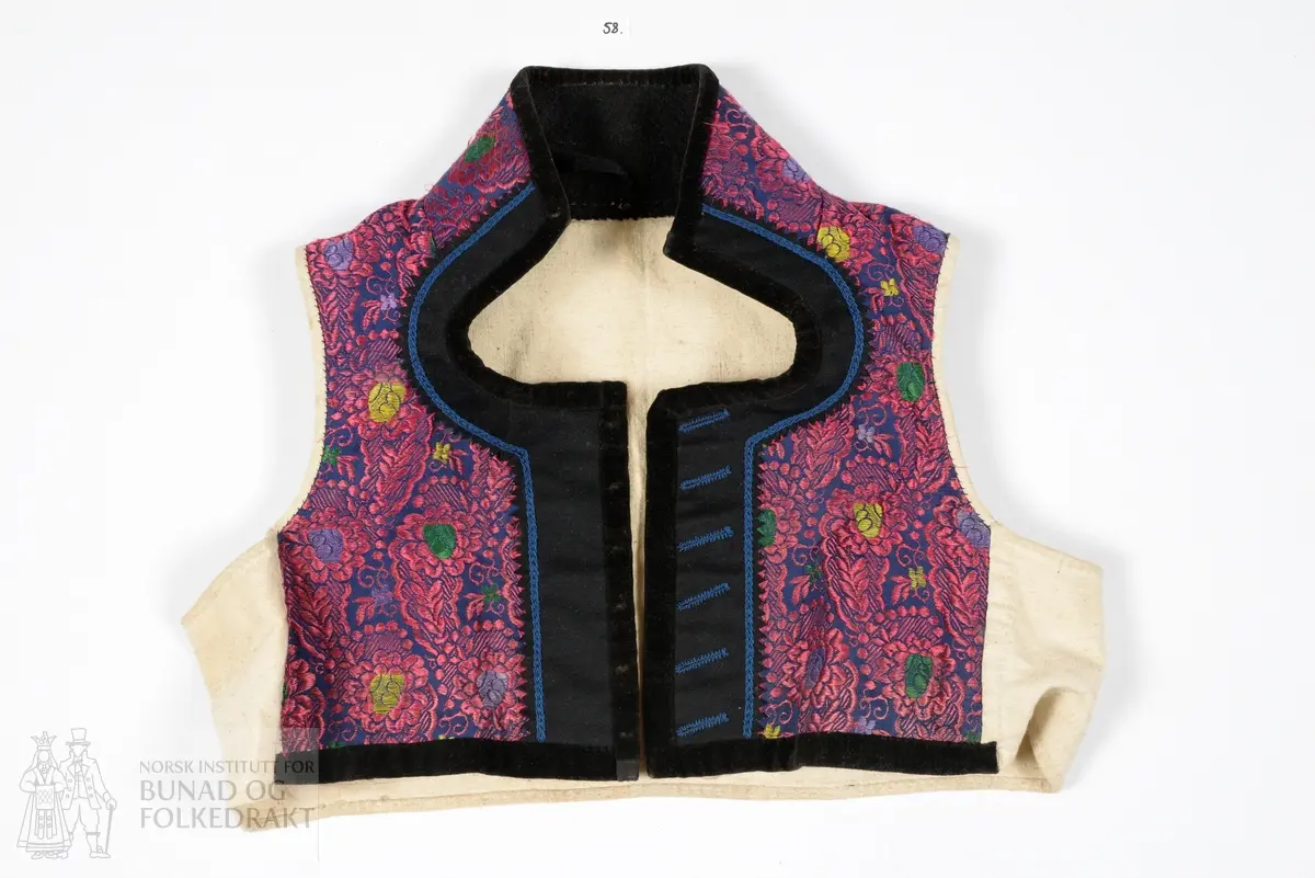 Vest, sauma av silkedroguet. Blå satengbotn med rosa flotterande trådar og innvove små blomster i ulike fargar. Framkant, krage og nedkant er påsydd ein smal svart kant av bomullsfløyel. Innanfor denne i framkant og krage er det påsydd eit breitt stykkje svart tynn fast klede. Denne er klipt med taggar etter sårkanten, pryda med tre tette konturstingsrader med blå ulltråd. 
Kragen er fora med tjukk brunsvart vadmål, sydd med to langsgåande avstivande stikningar. Ryggen er ufora, sauma av litt grov bomullslerret. 
Same type for i framstykkja. 
I ermholet på framstykkjet er foret bretta dobbelt rundt på framsida, og sydd ned med små javne faldesting. Sidesaumane er drage eit godt stykkje inn på kvart framstykkje. 
Eine framkanten har påsydd 6 stk skråstilte knapphol sauma av same type blå ulltråd. Merkjer etter knappefeste gjennom fløyelskanten på andre framstykkjet. 
I sidesaumen er hovudstoff framstykkje og ryggfor sydd saman med attersting, deretter er for framstykkjet bretta inn og sydd ned 1 cm bak den fyrste saumen, med faldesting som syner som prikk på retta.