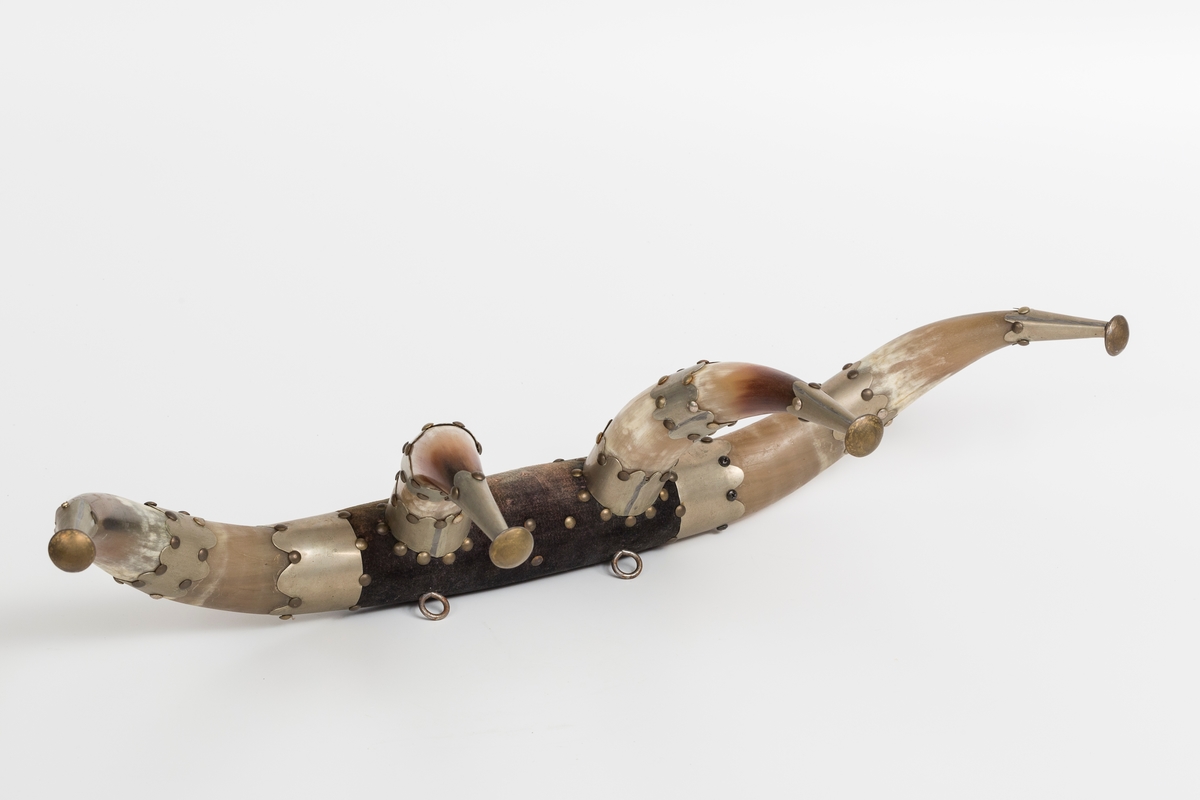 Knaggrekke antagelig håndlaget av kuhorn. 4 horn/kangger. Montert med "dekorative" beslag som er spikret og dekornaglet på hornene. Kledd med cordfløyelstoff på midtparti. To øyeskruer på toppen for feste til vegg.